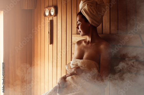 Young woman relaxing and sweating in hot sauna wrapped in towel. Girl In Sauna. Interior of Finnish sauna, classic wooden sauna with hot steam. Russian bathroom. Relax in hot sauna with steam