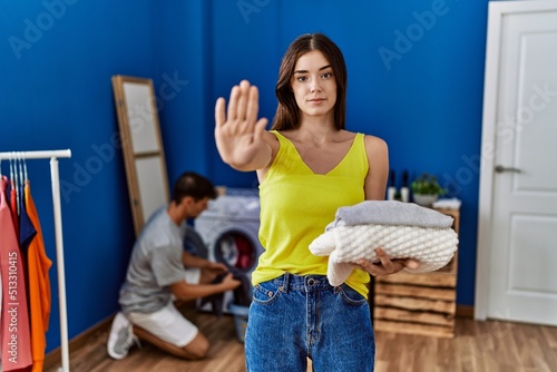 Young brunette woman holding folded laundry with open hand doing stop sign with serious and confident expression, defense gesture