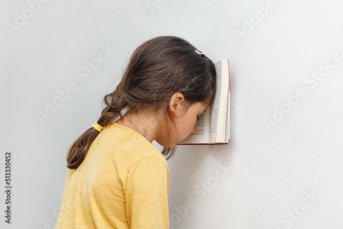 Sad and tired caucasian girl with dyslexia holds a book with her forehead. The child learns to speak and read correctly