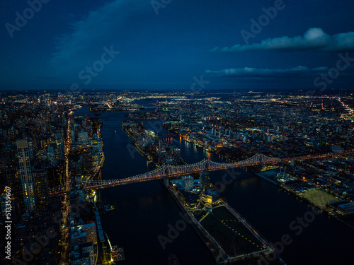 Aerial view of Queensboro Bridge and Roosevelt Island after sunset. Evening panoramic shot of city lights in urban boroughs. New York City, USA