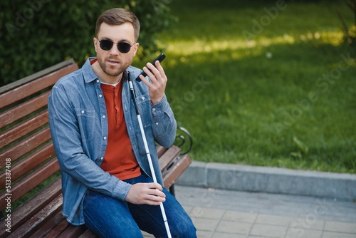 Visually impaired man with walking stick, sitting on bench in city park. Copy space