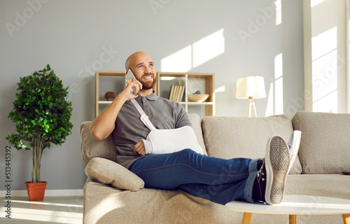 Relaxed man with broken arm having fun talking on phone with friends during rehabilitation at home. Cheerful bald young man in immobilizer on injured arm sitting on sofa throwing legs on coffee table.