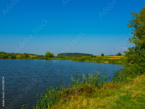 Rural lake in Ukraine in nice summer day. Quiet landscape in midday with blue water. Beautiful summer landscape at the bank of a pond with green trees.
