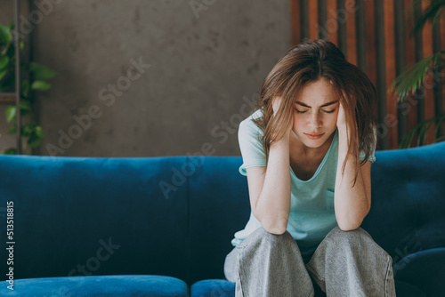 Young frustrated ill woman she 20s wear casual clothes mint t-shirt holding head suffer from headache close eyes sit on blue sofa indoor rest at home in own room apartment. People lifestyle concept