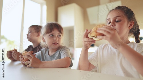 children eat burgers. fast food burger. fun a group of small children in the kitchen greedily eat fast food burgers. big family small kids having breakfast in the morning in the kitchen eating burger