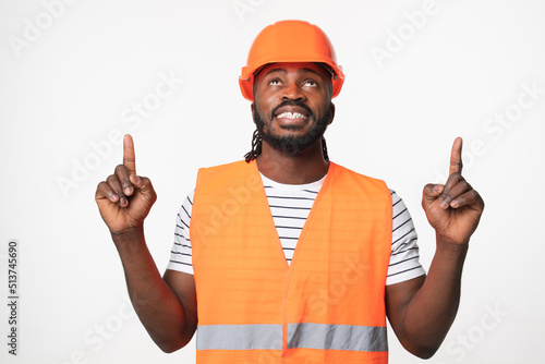 Young african-american man construction worker engineer architect in reflective orange vest and hardhat pointing at copy space upwards isolated in white background