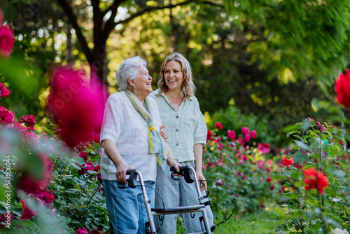 Adult granddaguhter supporting her senior grandmother when taking her for walk with walker in park in summer.