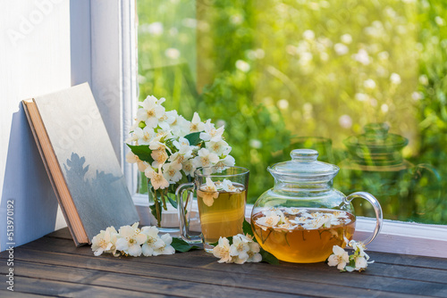 Hot herbal tea in glass teapot, cup and beautiful bouquet of jasmine flowers on windowsill at summer day near garden