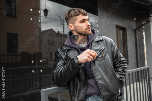 Handsome brutal hipster guy with a cigarette in fashion black outfit with leather jacket and hoodie walking in the city and smoking a cigarette