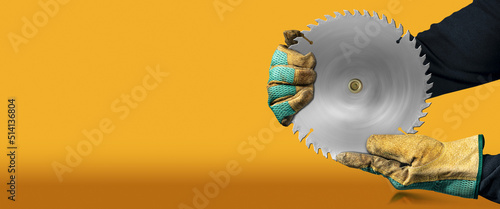 Carpenter with protective work gloves holding a metal circular saw blade, on orange and yellow background with copy space and reflections.