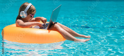 Little business man relaxing in the pool with laptop. Kid online working on laptop, swimming in a sunny turquoise water pool.