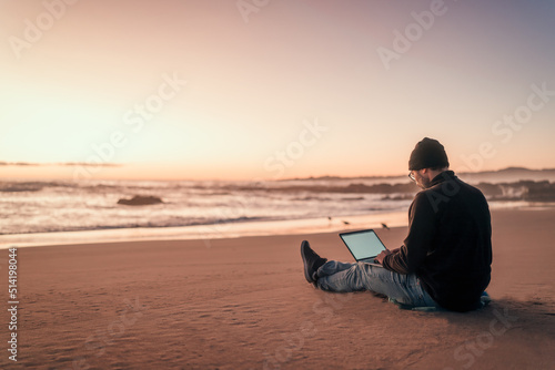 silhouette of a person working on his laptop outdoors on the beach at golden hour, back view.