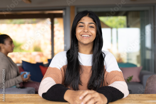 Portrait of happy biracial teenage girl sitting at table while mother with cup sitting in background