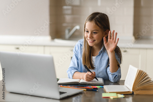 Online lesson, distance learning, homeschooling during quarantine. Caucasian pretty schoolgirl at online lesson by video call waves hand, greeting teacher, at home using laptop, sits at the work desk