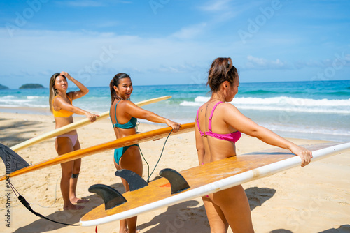 Group of Asian woman surfer in swimwear holding surfboard walking together on tropical beach in sunny day. Female friends enjoy outdoor activity lifestyle water sport surfing on summer vacation
