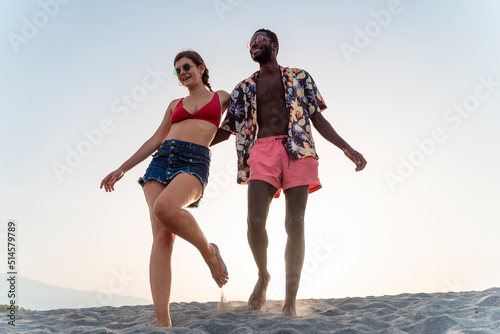 Engaged biracial couple happily walking barefoot in the sand holding together - multi-ethnic people relationship in the summer lifestyle concept