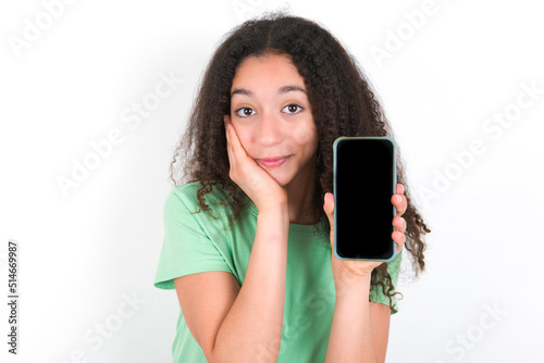 Teenager girl with afro hairstyle wearing green T-shirt over white wall hold hand modern technology use touch face palm astonished impressed scream wow omg unbelievable unexpected