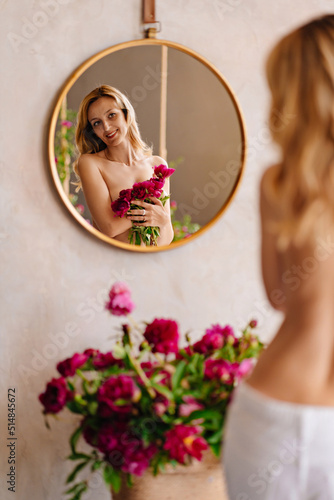a blonde woman with a bare back and a bouquet of peonies looks at in the mirror.