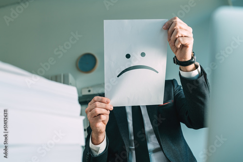 Unhappy businessman holding paper with frowning emoticon in office interior