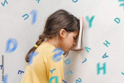 Sad and tired caucasian girl with dyslexia holds a book on her forehead. Flying tangled letters in the air. The child learns to speak and read correctly