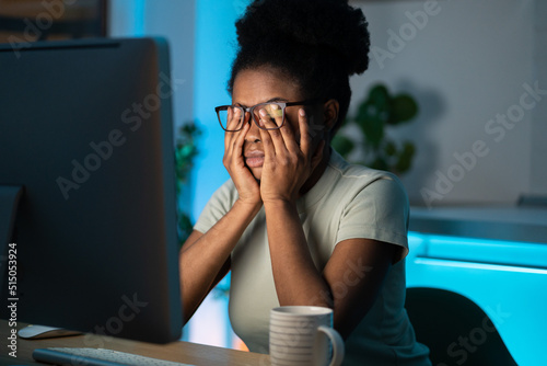 Symptoms of overworking. Young tired overworked african woman freelancer in eyeglasses working late from home, sitting in front of monitor, feeling eye strain and fatigue during computer work. 