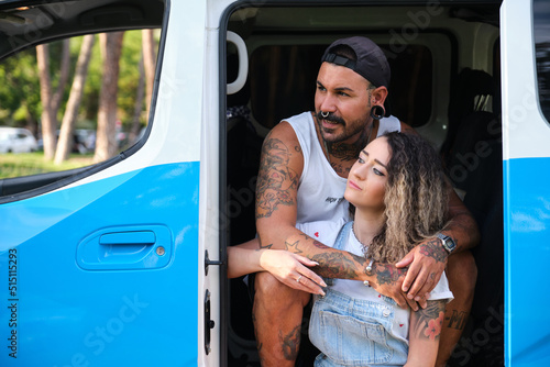 Young tattooed couple hugging and looking at a side, sitting in the side of the van.