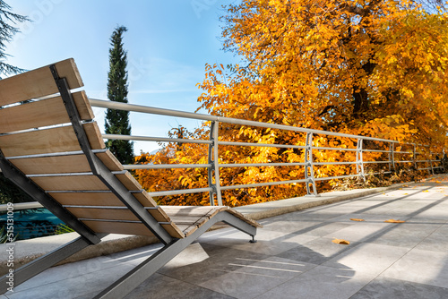 Close view of modern wooden empty lounger chair on patio balcony terrace against bright yellow orange autumn tree and blue sky. Fall season vacation and travel concept