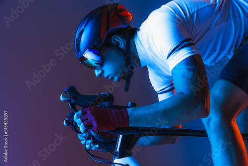 Closeup portrait of professional male cyclist in sports uniform, goggles and helmet on blue background in neon. Concept of sport fashion, race, competition