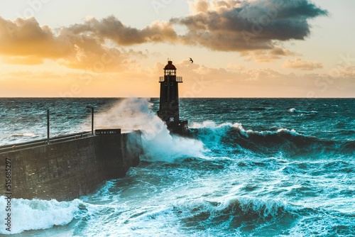 Large waves crash against the stone tower of the lighthouse at high tide at sunset