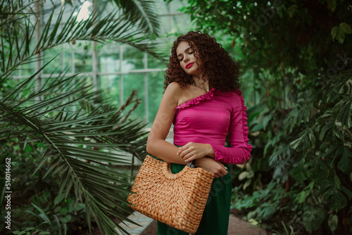 Fashionable curly woman wearing pink asymmetric ruffled blouse, holding straw wicker top handle bag, posing in tropical garden. Summer fashion, lifestyle conception. Copy, empty space for text 