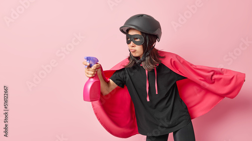Hygiene cleaning concept. Serious woman imagines herself being superhero wears cape and helmet holds detergent looks at dirty room readty to save you from dirt isolated over pink background.