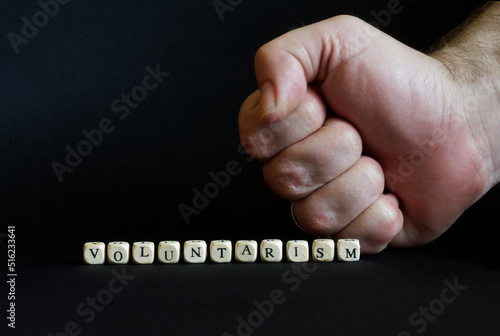 Inscription voluntarism and a clenched male fist on a black background. The concept of voluntaristic oppression, coercion and lack of freedom.