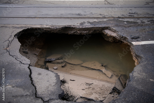 Huge sinkhole on busy asphalt road surface on which cars drive. Accident situation on a city street due to cracks in asphalt. Broken hole filled with muddy water.