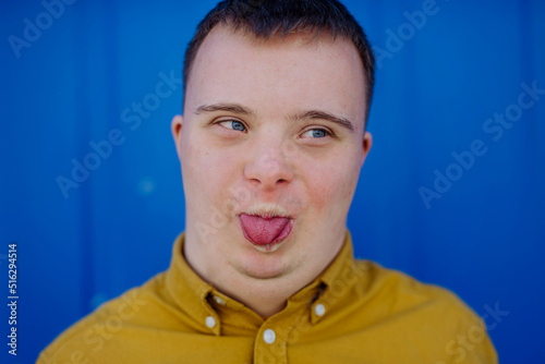 Close-up of happy young man with Down syndrome grimacing and sticking out tongue against blue background.