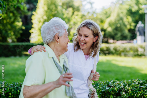 Portrait of caregiver with senior woman on walk in park with shopping bag, looking at caemra.