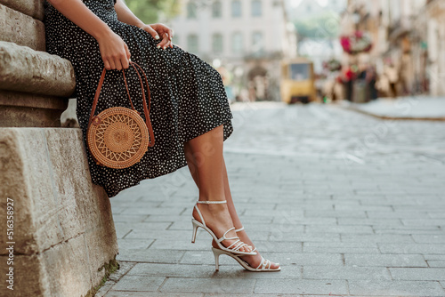 Trendy summer rotan wicker bag, white strap sandals in stylish female outfit. Woman posing in street of European city. Fashion details. Copy, empty space for text