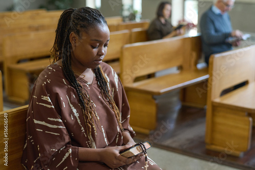 Young African American woman holding Bible and rosary beads while sitting on bench in evangelical church during sermon
