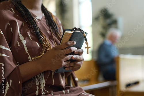 Hands of young black woman with Holy Bible and rosary beads with small wooden cross standing in church and praying after sermon