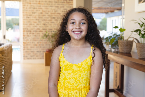 Portrait of smiling biracial cute girl with long curly hair wearing yellow dress standing at home