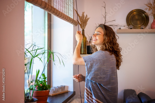 Beautiful happy young woman opening rolled bamboo blinds in country house in summer morning