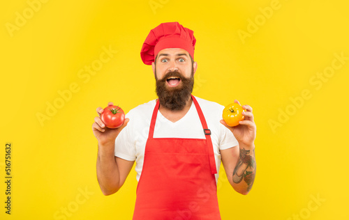 Surprised man in cooking apron and toque holding red and yellow tomatoes yellow background, cook