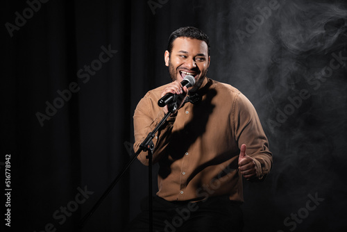 happy indian comedian sitting on chair and performing stand up comedy into microphone on black with smoke.