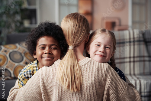 Portrait of happy adoptive children looking at camera while embracing their foster mother at home