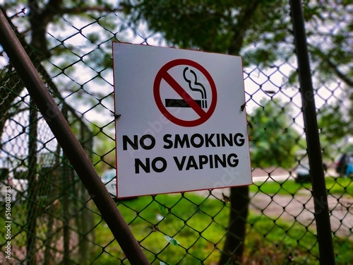 Close up of a no smoking, no vaping sign fixed on metal chain fence in front of school playground and entry area.