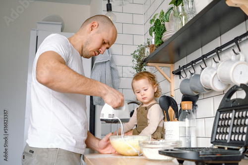 A dad and baby toddler cooking together. A father and kid have fun spending time together on the kitchen baking waffles at home. A man mixing ingredients for dough using blender.