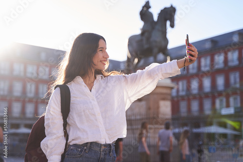 Happy caucasian woman is taking a selfie smiling at the camera in front of the Equestrian Monument to King Felipe III of Spain in the Plaza Mayor in Madrid