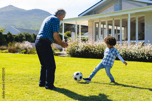 Caucasian grandfather playing soccer with biracial grandson on grassy land in yard on sunny day