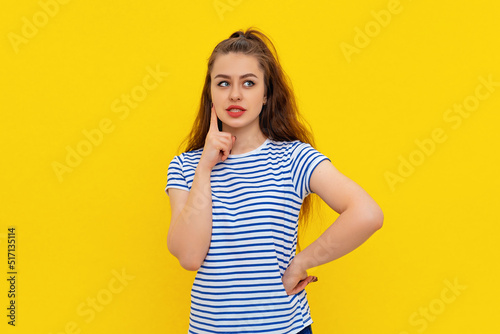 Image of girl with brown-haired hair thinking, posing thoughtful, making choice or decision, looking aside, standing in white-blue striped t shirt over yellow background