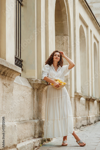 Beautiful curly brunette woman wearing trendy summer white linen shirt, long skirt, sandals, holding bowl with lemons, posing in street of European city. Full-length outdoor portrait