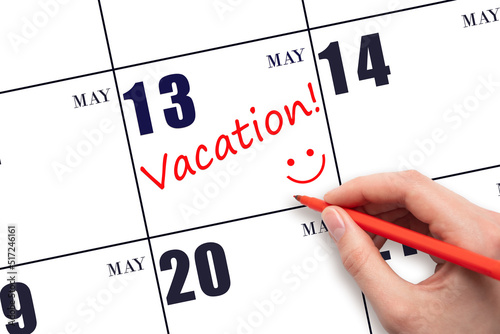 A hand writing a VACATION text and drawing a smiling face on a calendar date 13 May. Vacation planning concept.
